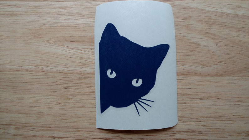 May include: A navy blue cat silhouette peeking from the side, with white eyes and whiskers, on a white sticker. The sticker is placed on a light brown wooden surface.