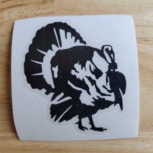 May include: A black turkey silhouette sticker on a white square background. The turkey is depicted in profile, with its head and tail feathers prominently displayed. The sticker is likely for decoration or crafting.