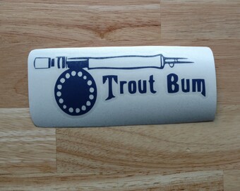 May include: A rectangular sticker with a fishing rod illustration and the words "Trout Bum" in navy blue. The fishing rod is light blue, and the reel has a navy blue circular design. The sticker is on a wooden surface.