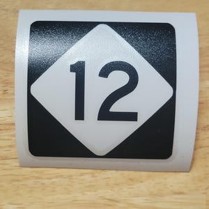 Op de afbeelding: Een rechthoekige sticker met een witte ruit met het zwarte nummer "12" op een zwart vierkant. De sticker heeft een witte rand en ligt op een houten oppervlak.