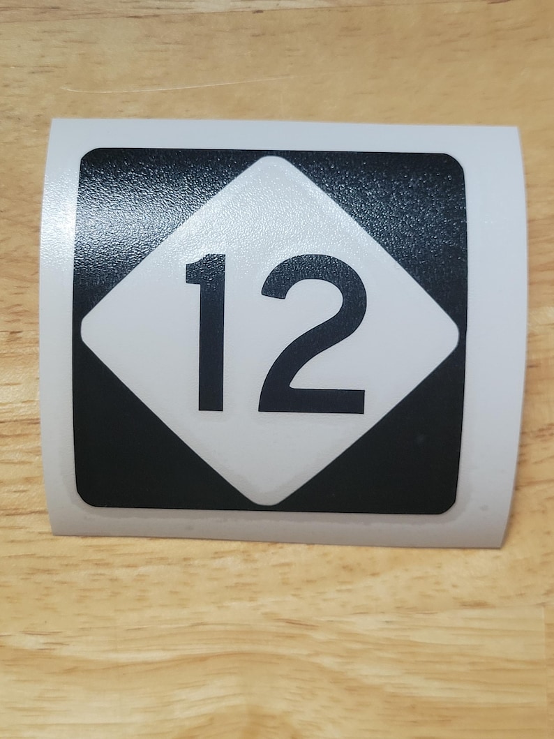 Op de afbeelding: Een rechthoekige sticker met een witte ruit met het zwarte nummer "12" op een zwart vierkant. De sticker heeft een witte rand en ligt op een houten oppervlak.