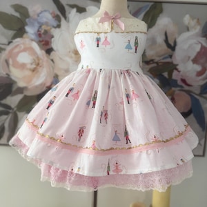 May include: A sleeveless dress featuring a white bodice with a small pink bow and a light pink skirt. The fabric is printed with small illustrations of nutcracker figures and ballerinas. The skirt has gold trim and a light pink lace hem.