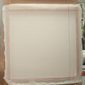 May include: A rectangular, off-white fabric stretched over a wooden frame. The fabric is secured with a pink-lined border, and the frame is supported by a brown cardboard backing. The setup is likely for a craft project.