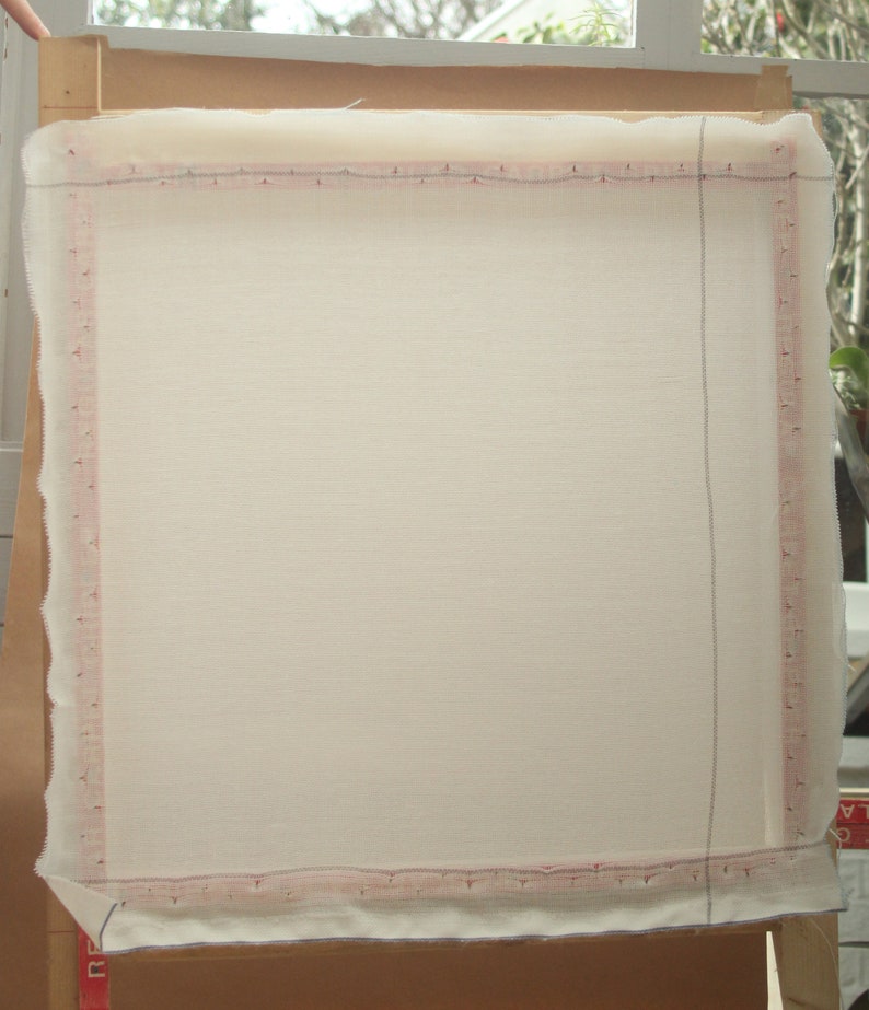 May include: A rectangular, off-white fabric stretched over a wooden frame. The fabric is secured with a pink-lined border, and the frame is supported by a brown cardboard backing. The setup is likely for a craft project.