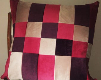 May include: A square decorative pillow with a patchwork design. The pillow features a grid pattern of various colors, including shades of pink, purple, and beige. The pillow is resting on a wooden chair.