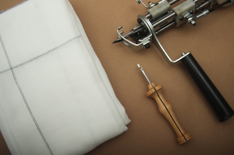 May include: A white rug hooking fabric with a gray grid pattern is next to a rug tufting gun and a wooden handle punch needle tool. The tools are on a brown surface.