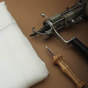 May include: A white fabric square, a tufting gun, and a wooden handle punch needle are arranged on a brown surface. The tufting gun is silver and black. The punch needle has a silver needle and a light brown wooden handle. The fabric is white.