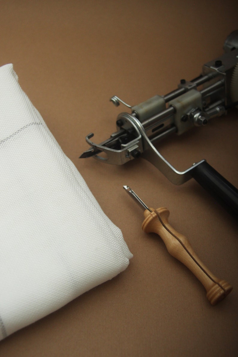 May include: A white fabric square, a tufting gun, and a wooden handle punch needle are arranged on a brown surface. The tufting gun is silver and black. The punch needle has a silver needle and a light brown wooden handle. The fabric is white.
