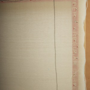 May include: Close-up of a cream-colored rug canvas stretched over a wooden frame. The canvas has a grid pattern with red and gray lines along the edges. The edges of the canvas are slightly frayed.