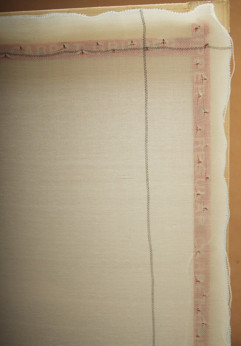 May include: Close-up of a cream-colored rug canvas stretched over a wooden frame. The canvas has a grid pattern with red and gray lines along the edges. The edges of the canvas are slightly frayed.