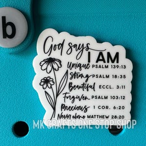 Op de afbeelding: Witte sticker met zwarte tekst en een bloemmotief. De tekst luidt "God zegt dat ik ben Uniek Psalm 139:13 Sterk Psalm 18:35 Mooi Eccl. 3:11 Vergeven Psalm 103:12 Kostbaar 1 Cor. 6:20 Nooit alleen Mattheüs 28:20".
