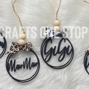 May include: Four black wooden ornaments with a white and brown leopard print bow. Each ornament has a different name written on it: Mom, Mimi, Gigi, and Nana.
