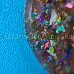 Pu&ograve; includere: Primo piano di un oggetto in resina con pezzi di glitter iridescenti in vari colori, tra cui rosa, verde e oro. L'oggetto &egrave; posto su uno sfondo blu brillante. Il testo "MKCrafts OneStop Shop" &egrave; visibile.
