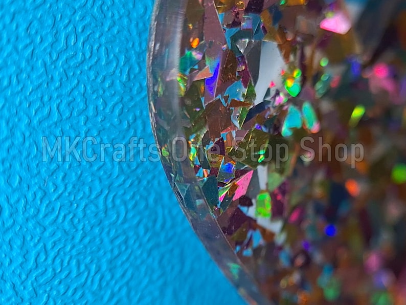 Pu&ograve; includere: Primo piano di un oggetto in resina con pezzi di glitter iridescenti in vari colori, tra cui rosa, verde e oro. L'oggetto &egrave; posto su uno sfondo blu brillante. Il testo "MKCrafts OneStop Shop" &egrave; visibile.