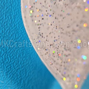 Pu&ograve; includere: Primo piano di un oggetto trasparente a forma di cuore pieno di glitter argentati e macchie iridescenti. L'oggetto &egrave; posto su uno sfondo blu strutturato. Il testo "MKCrafts One-Stop Shop" &egrave; visibile.