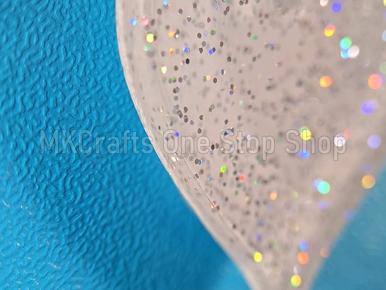 Pu&ograve; includere: Primo piano di un oggetto trasparente a forma di cuore pieno di glitter argentati e macchie iridescenti. L'oggetto &egrave; posto su uno sfondo blu strutturato. Il testo "MKCrafts One-Stop Shop" &egrave; visibile.