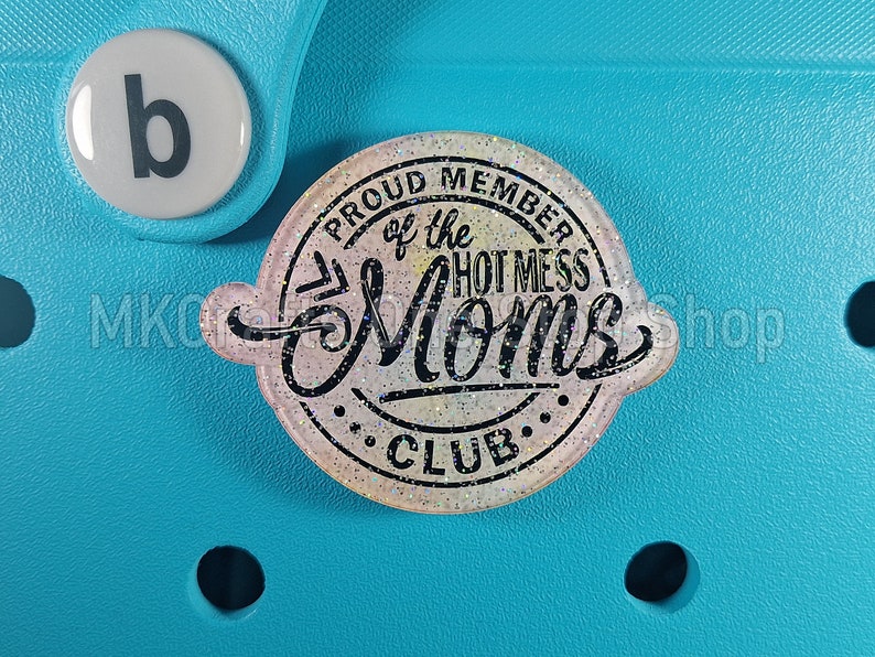 K&ouml;nnte beinhalten: Ein wei&szlig;er und rosafarbener Glitzer-Anh&auml;nger mit dem Text "Proud Member of the Hot Mess Moms Club" in schwarzen Buchstaben.