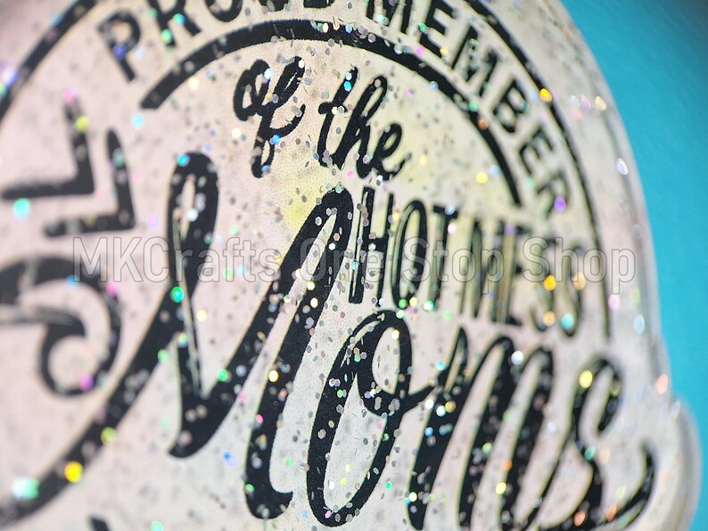 K&ouml;nnte beinhalten: Nahaufnahme eines wei&szlig;en, glitzernden Aufklebers mit schwarzem Text, der "Proud Member of the Hottest Moms" lautet.