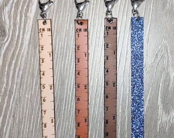 May include: Four ruler-shaped keychains. Three are leather in tan, brown, and dark brown, with inch and centimeter markings. The fourth is blue glitter. Each has a silver clasp. The rulers measure 9 inches (22.86 cm) long.