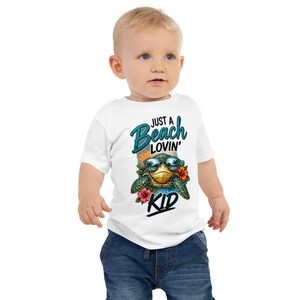 May include: A white short-sleeved t-shirt featuring a graphic of a sea turtle wearing sunglasses, surrounded by tropical flowers and palm trees. The text "JUST A Beach LOVIN' KID" is printed in blue and black lettering above and below the turtle.