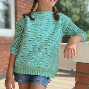 Può includere: Una giovane persona indossa una maglia a uncinetto blu chiaro con un motivo a macchie verdi e blu. La maglia ha un collo a barca e maniche a tre quarti. La persona è in piedi su un portico in legno con un pilastro di mattoni dietro di lei.