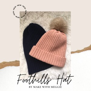 May include: Two crocheted hats, one navy blue and one light pink with a fluffy pom pom. The hats are on a white fluffy surface. The text "Foothills Hat" and "By Make With Meggie" is below the hats.