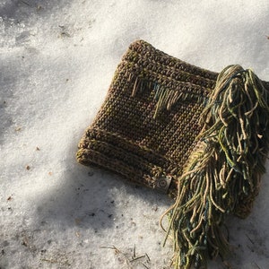 Puede incluir: Una bufanda de crochet verde y marrón con una larga franja. La bufanda está sobre una superficie nevada.