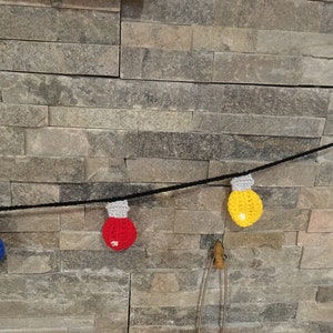 May include: A string of crocheted light bulbs in red, blue, yellow, and green hanging on a stone wall.