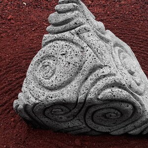 May include: A gray, porous stone sculpture shaped like a triangular pyramid. Each of the three visible faces features a deeply carved, swirling spiral pattern that extends from the center toward the edges of the textured, volcanic-like rock surface.