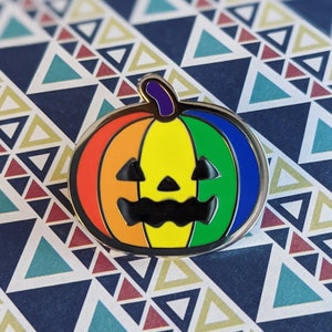 May include: A rainbow-colored enamel pin shaped like a jack-o'-lantern. The pumpkin is outlined in silver and has a black stem and facial features.