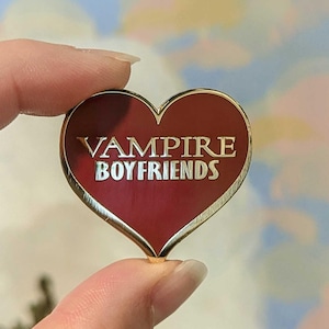 Pode incluir: Um alfinete vermelho em forma de coração com borda dourada e o texto "Vampire Boyfriends" em letras douradas.