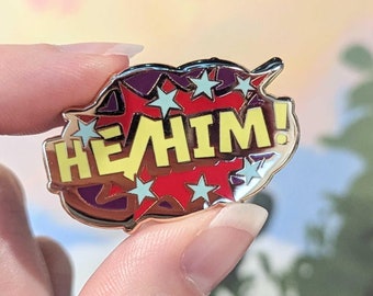 May include: A gold-colored enamel pin with a red and purple comic book style speech bubble. The speech bubble says "HE HIM!" in yellow letters with white stars around it.