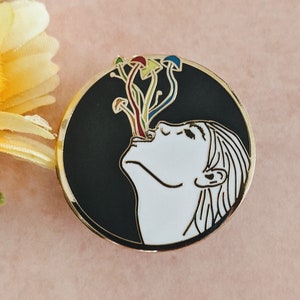 Op de afbeelding: Een zwart-witte emaille pin met een persoon met zijn mond open, die kleurrijke paddenstoelen inademt. De pin is rond en heeft een gouden rand.