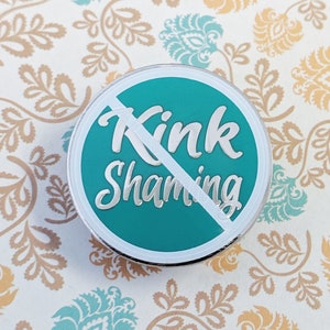 May include: A teal blue enamel pin with a white circle and diagonal line through it. The text "Kink Shaming" is written in silver lettering.