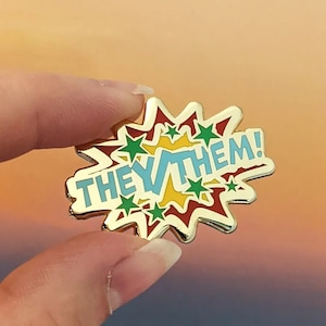 以下が含まれることがあります： 漫画風の吹き出しデザインの金色のピン。吹き出しには「THEY/THEM!」という青い文字が黄色と赤のアクセントで書かれています。吹き出しは赤、黄、緑の星で囲まれています。