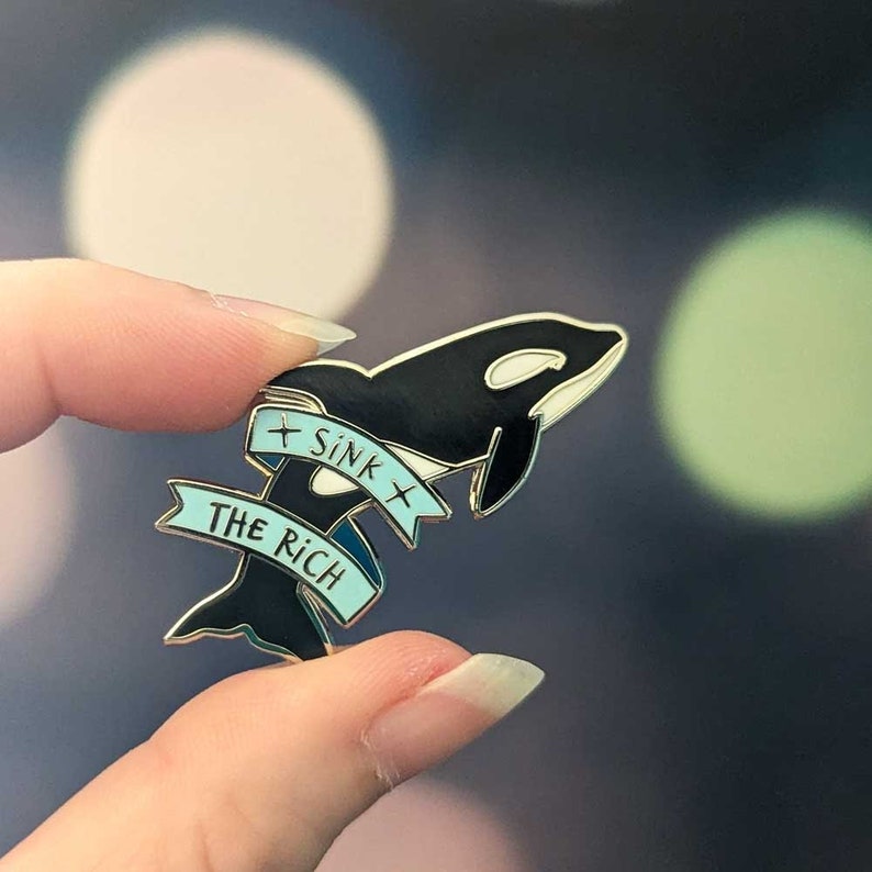 An enamel pin shaped like an orca whale jumping through a banner that says "Sink the rich"