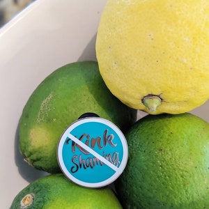 May include: A teal blue enamel pin with a white slash through the words "Kink Shaming". The pin is sitting on top of limes.