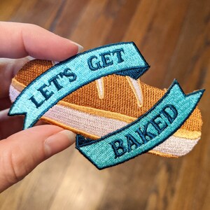 May include: A blue and white embroidered patch with a loaf of bread and the text "Let's Get Baked".