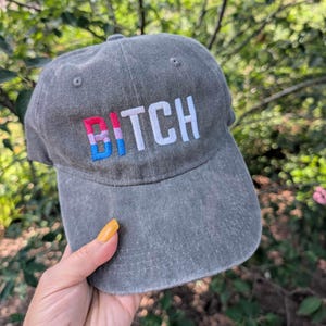 Peut inclure: Une casquette de baseball gris anthracite avec un motif brodé sur le devant. Le mot BITCH est cousu en fil blanc, la lettre B présentant un motif à rayures verticales dans des tons rose, violet et bleu. La casquette a une visière courbée.