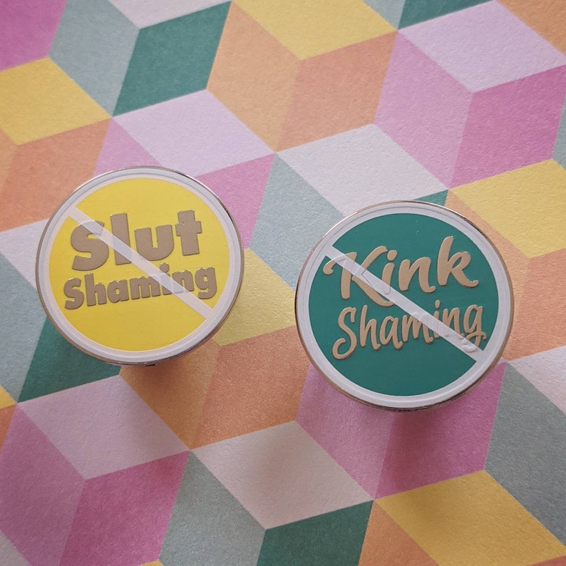 May include: Two round pins with a white slash through the center. The left pin is yellow with the text "Slut Shaming" in gold lettering. The right pin is green with the text "Kink Shaming" in gold lettering.