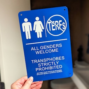May include: A blue rectangular sign with white text and graphics. The sign features stick figures representing male and female genders, a circle with "TERFS" crossed out, and the phrases "ALL GENDERS WELCOME" and "TRANSPHOBES STRICTLY PROHIBITED."