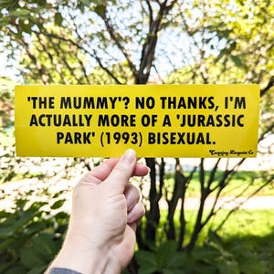 May include: Yellow bumper sticker with black text that reads: 'THE MUMMY'? NO THANKS, I'M ACTUALLY MORE OF A 'JURASSIC PARK' (1993) BISEXUAL.  Cunning Linguist Co