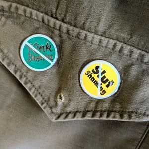May include: Two round pins with a white slash through the center. The top pin is green with the text "Kink Shaming" in black. The bottom pin is yellow with the text "Slut Shaming" in black.