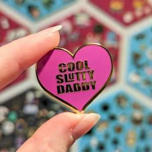 May include: A heart-shaped enamel pin with a bright pink background and gold trim. The pin features the words "COOL SLUTTY DADDY" in gold lettering. The pin is held up by a person's fingers.