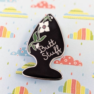 Könnte beinhalten: Schwarzer Emaille-Pin mit einer weißen Blume und grünen Blättern. Der Pin trägt die Aufschrift "Butt Stuff" in weißen Buchstaben.