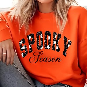 Puede incluir: Una sudadera naranja con las palabras "SPOOKY Season" en letras negras y un estampado de fantasmas. La sudadera es de cuello redondo y está hecha de un material suave. Ideal para Halloween.