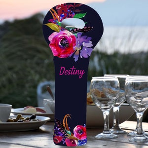 May include: A navy blue wine bottle bag with a floral design featuring pink and purple flowers, green leaves, and feathers. The word "Destiny" is written in pink script. The bag is designed to hold a wine bottle.