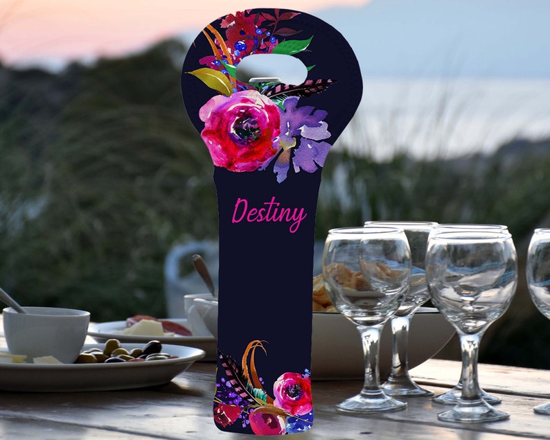 May include: A navy blue wine bottle bag with a floral design featuring pink and purple flowers, green leaves, and feathers. The word "Destiny" is written in pink script. The bag is designed to hold a wine bottle.