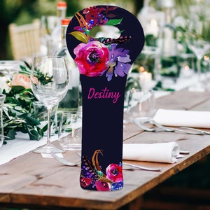 May include: A navy blue wine bottle bag with a floral design featuring pink and purple flowers, and the word "Destiny" in pink script. The bag has a handle and is decorated with feathers.