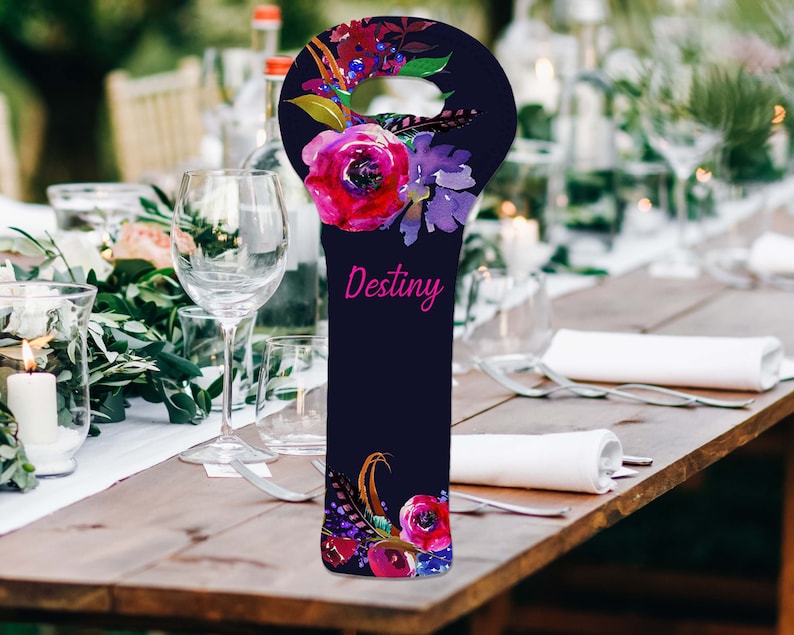 May include: A navy blue wine bottle bag with a floral design featuring pink and purple flowers, and the word "Destiny" in pink script. The bag has a handle and is decorated with feathers.